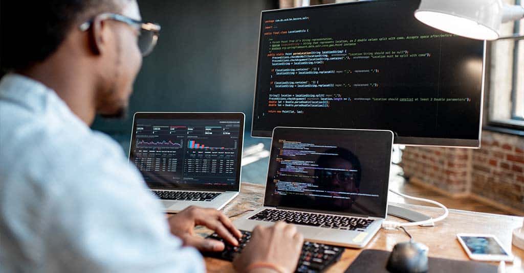 A man coding in front of three computer screens on his desk.