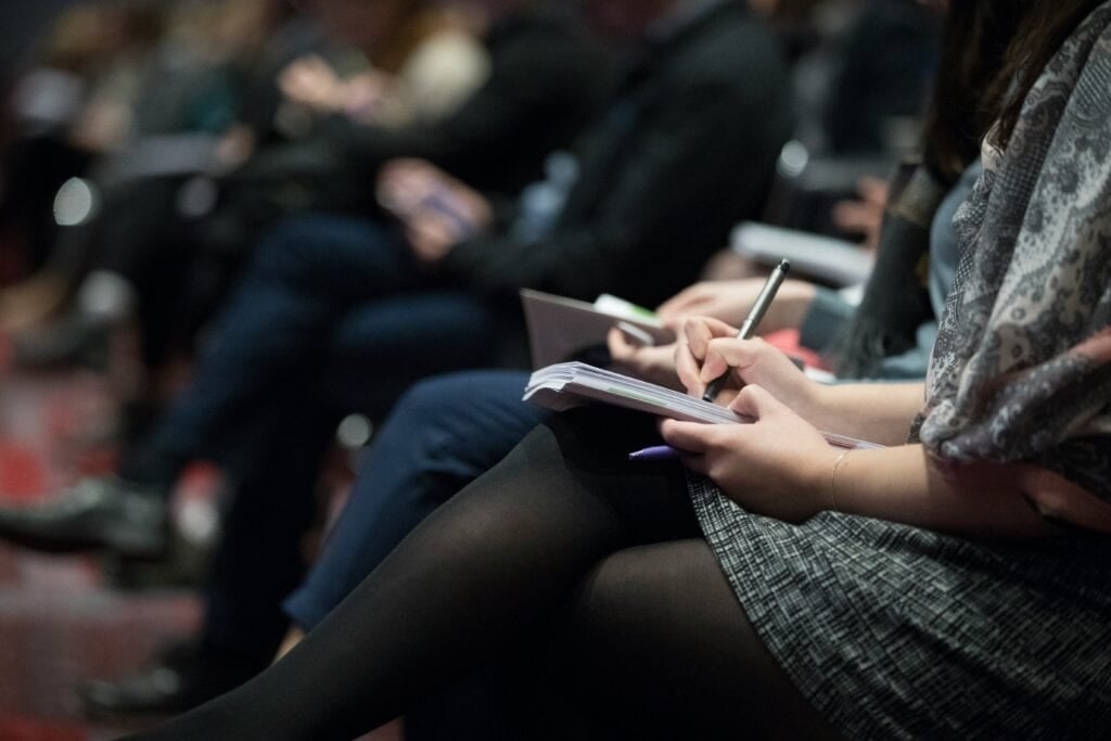 A close-up shot of several people in an auditorium taking notes on their notepads.